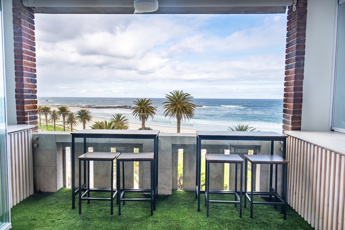 A balcony area features two high-top tables with accompanying stools, set against a backdrop of palm trees and ocean waves. Natural light illuminates the space, which is bordered by stone walls and partial glass panels that enhance the view of the beach.