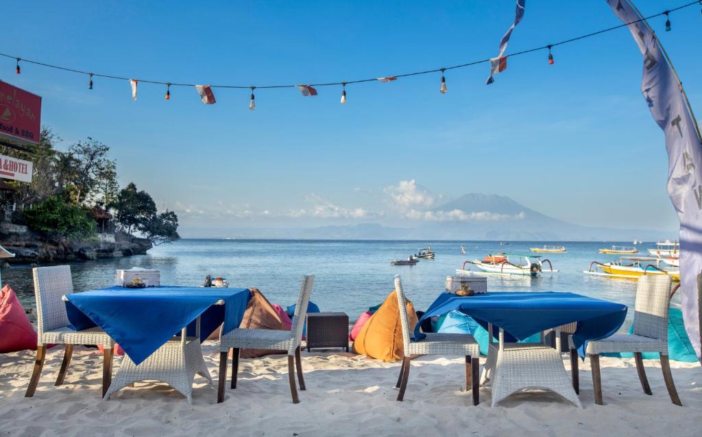 A selection of dining tables with blue tablecloths is arranged on the sandy beach, complemented by colorful bean bags nearby. Strings of lights are hung above, creating an inviting atmosphere. The serene water and distant mountains provide a picturesque backdrop.