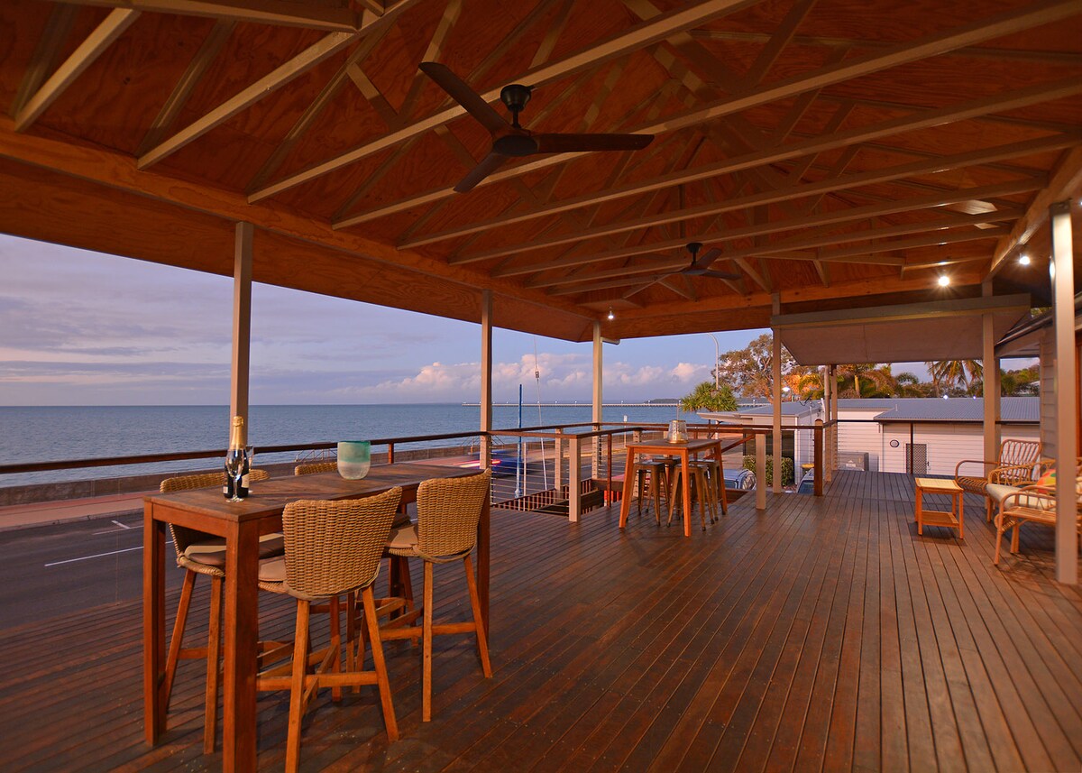A spacious deck is viewed overlooking the ocean, featuring wooden flooring with a natural finish and a wooden ceiling. Bar stools and a dining table are positioned near the railing, providing a perfect spot for enjoying the sea breeze and stunning water views.