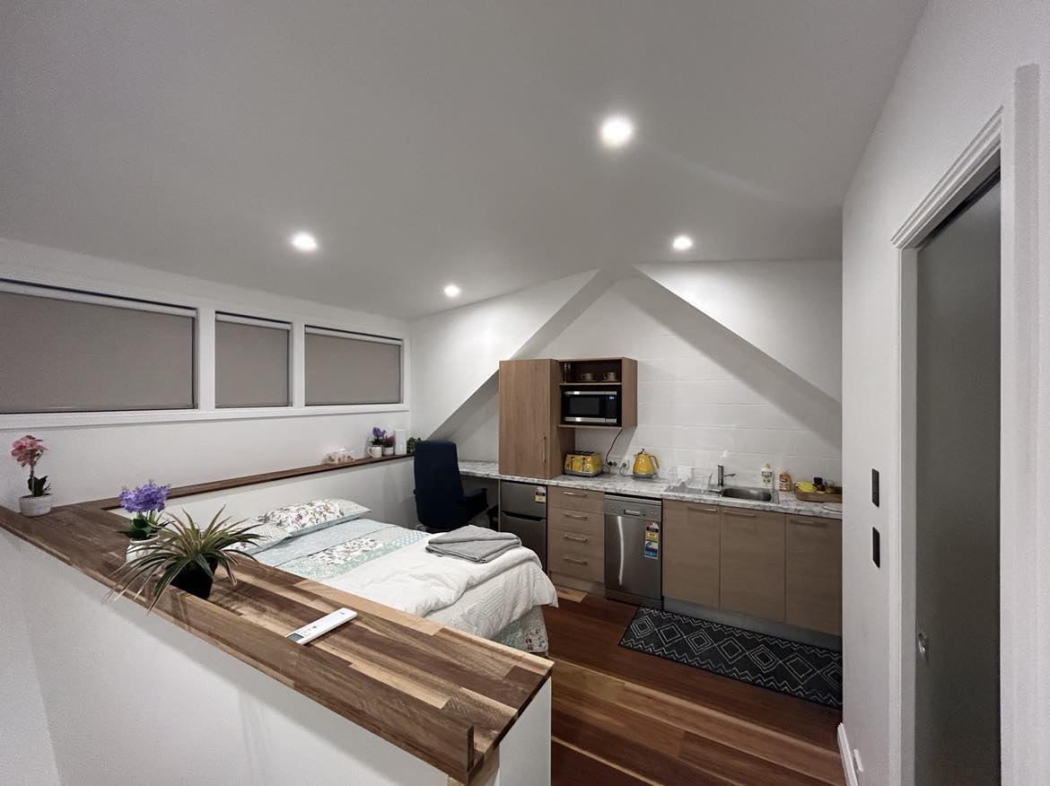 A well-organized studio apartment is presented, featuring a spacious bed with soft linens, a fully equipped kitchen with modern appliances, and a dedicated study area. Natural lighting is maximized through large windows above the kitchen, illuminating warm wooden floors and minimalist decor.