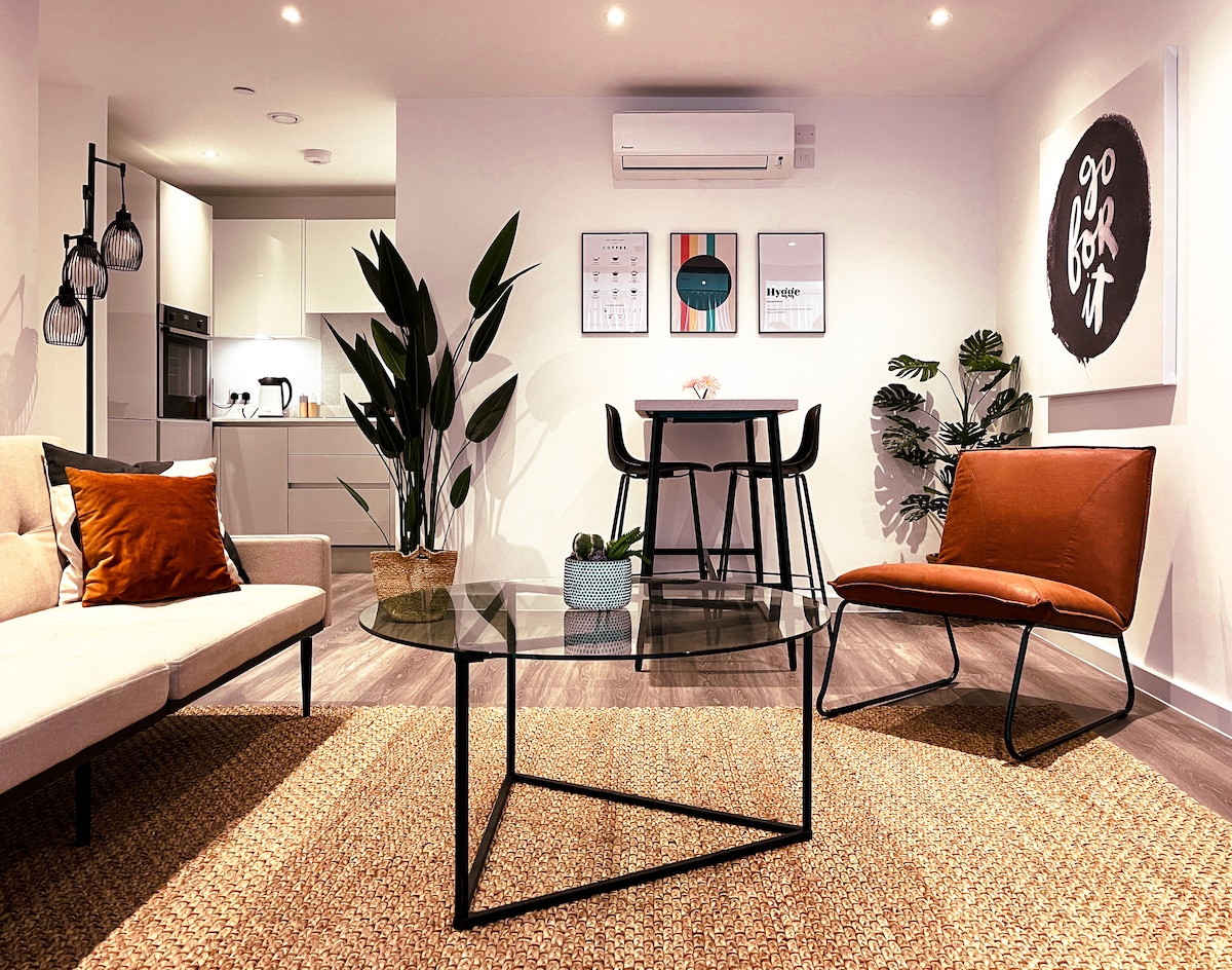 An open-plan living area features a stylish sofa and accent chairs, with a glass coffee table at the center. A dining table is set for four near the kitchen, which is partially visible. Green plants adorn the space, complementing the modern artwork on the walls.