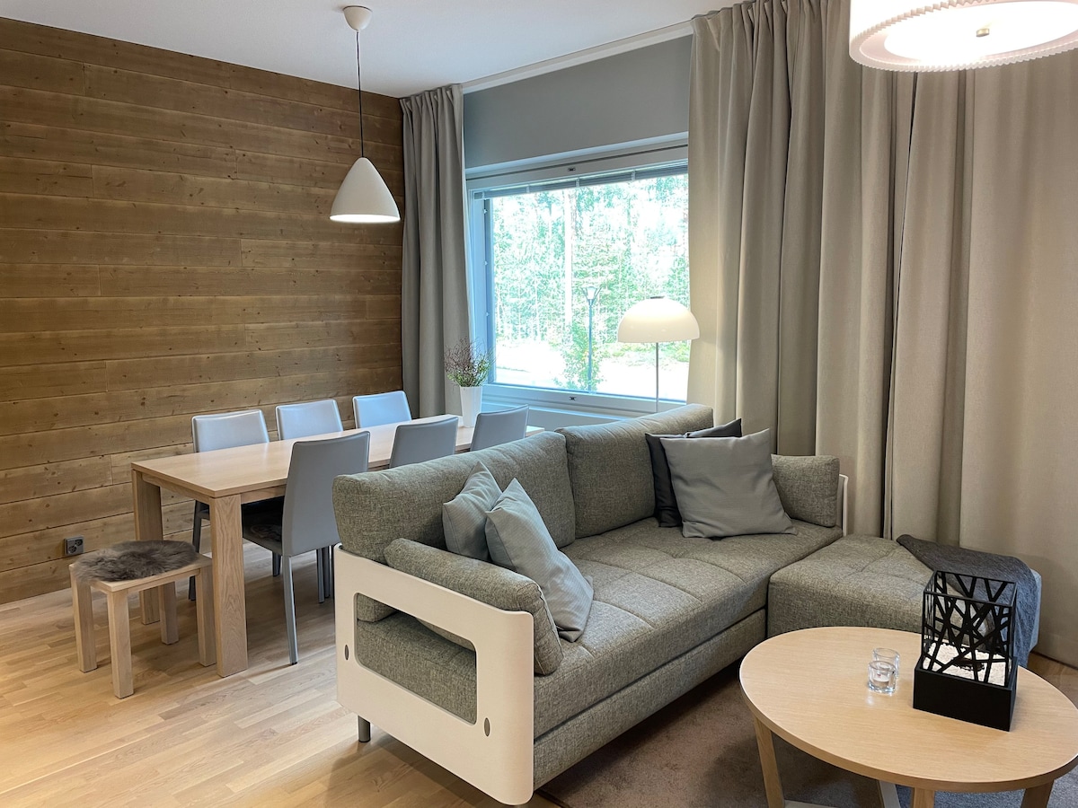 A light-filled living area is characterized by a comfortable corner sofa and a round coffee table. A dining table with six chairs sits adjacent. Large windows allow views of the surrounding greenery, while soft curtains frame the light wood paneling and flooring.