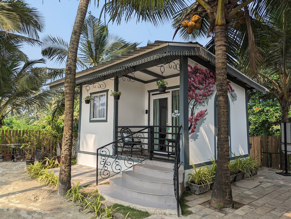 A quaint cottage is surrounded by lush palm trees and vibrant greenery. The entrance features a small stairway leading to a covered porch adorned with potted plants and a bright flower arrangement, inviting guests to enjoy the serene outdoor space.