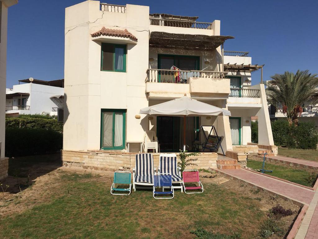 A two-story building features a covered balcony and outdoor seating area with colorful beach loungers. Lush green grass surrounds the property, and palm trees are positioned nearby, contributing to the space's relaxed atmosphere. Bright blue skies are visible above.
