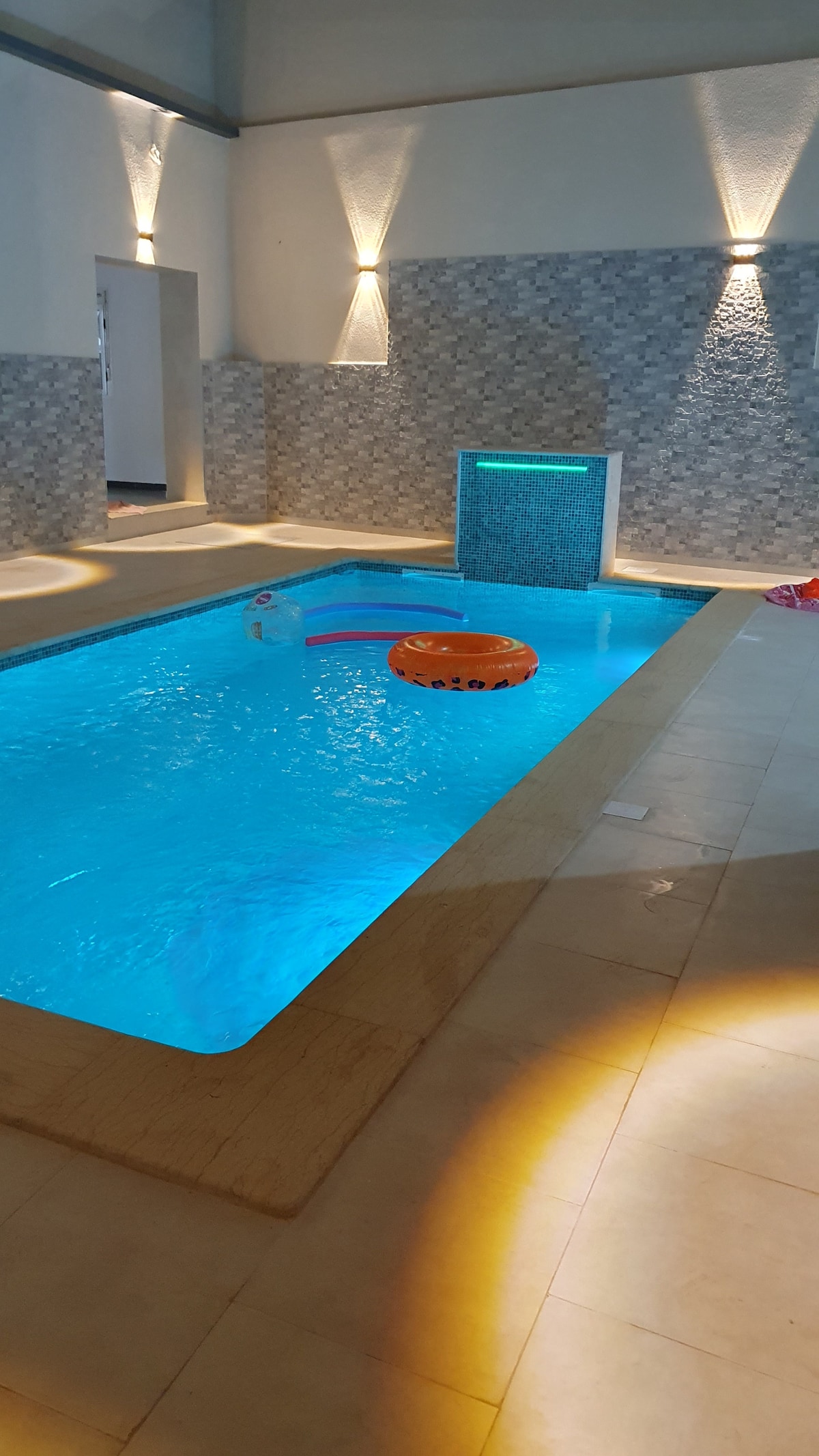 A private indoor swimming pool is highlighted by soft lighting and a decorative stone wall. The clear blue water reflects the light, with a yellow inflatable ring floating. The surrounding area features smooth tile flooring, creating a serene atmosphere for relaxation.