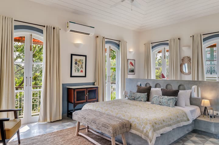 ✨Framed by arched French windows and soft linen curtains, this airy bedroom is an elegant mix of Mediterranean calm and Indo-Portuguese charm✨