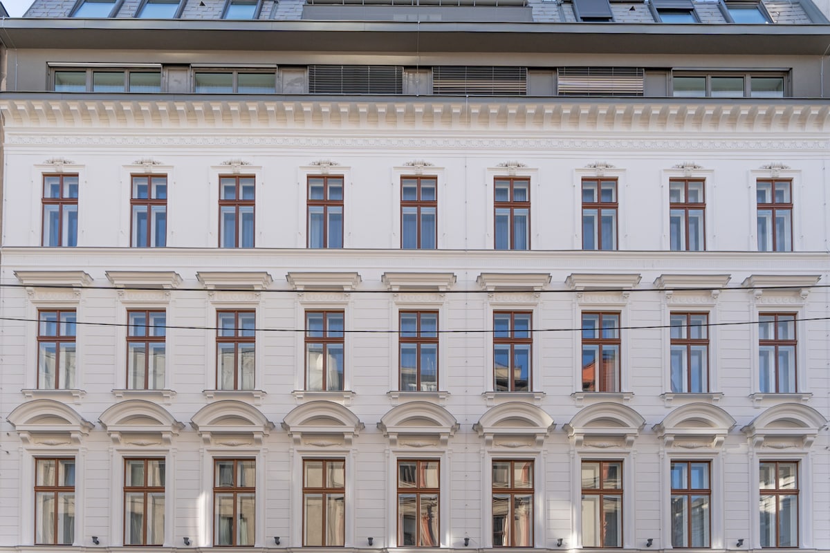 The exterior of the building features a classic architectural design with a white facade and multiple brown-framed windows. Decorative elements are present above the windows, contributing to the building's elegant appearance. The structure is several stories tall, showcasing a blend of traditional and modern design.