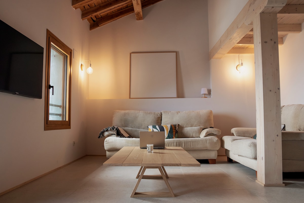 A welcoming living area features a light-colored sofa and an armchair positioned around a central wooden coffee table. Natural light fills the space through a window, highlighting exposed wooden beams and neutral wall tones. A television is mounted on the wall opposite the seating.