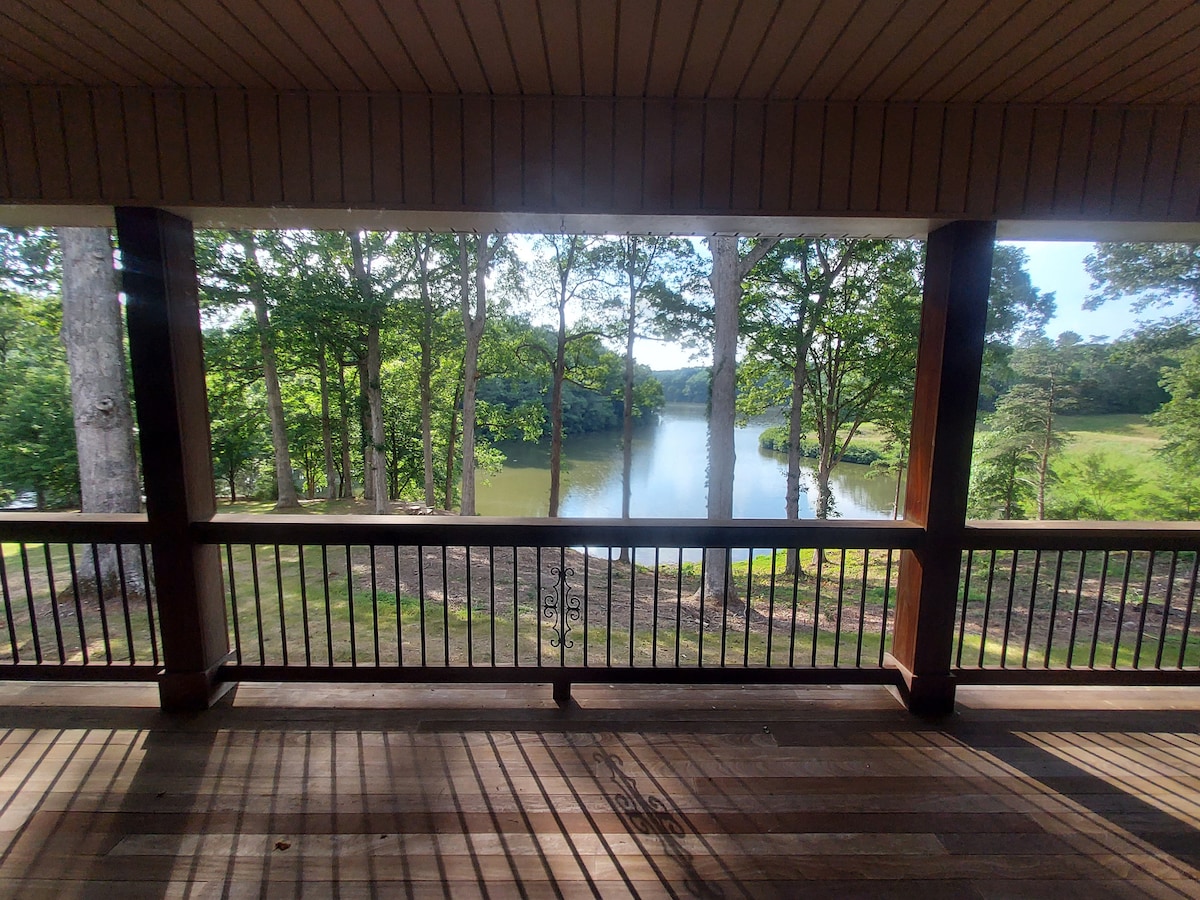A panoramic view is captured from a spacious wraparound porch, showcasing well-maintained trees and a serene lake. Natural light filters through the wooden beams, creating a harmonious blend of nature and architecture.