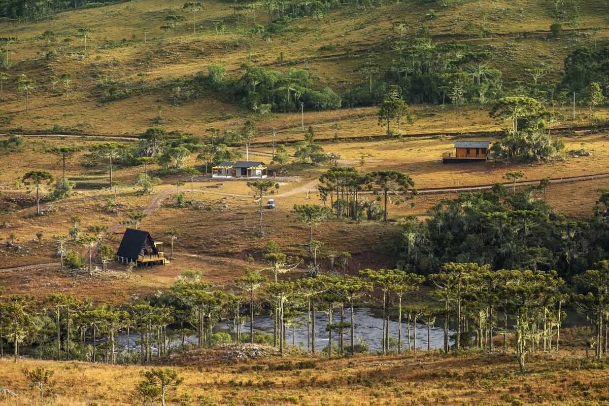 A panoramic view of the River House property shows rolling hills covered in greenery, interspersed with trees. Several buildings are positioned throughout the landscape, with a serene body of water visible in the foreground, reflecting the natural beauty of the surroundings.