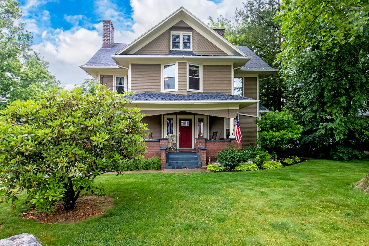 A historic home featuring a welcoming front porch with an American flag. Lush green landscaping surrounds the property, complemented by a symmetrically manicured lawn. Large trees provide shade, enhancing the tranquil setting of this inviting second-floor queen room at Casa Mirabella Guest House.
