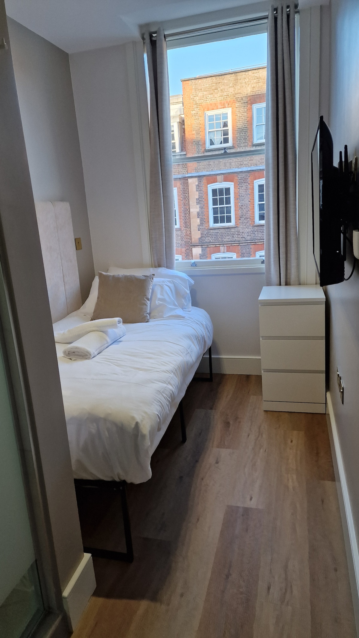 A compact room features a single bed dressed in crisp white linens and two neatly arranged towels. A tall chest of drawers stands next to the bed, and a wall-mounted smart TV is positioned opposite. Natural light floods the space through a tall window with light curtains.