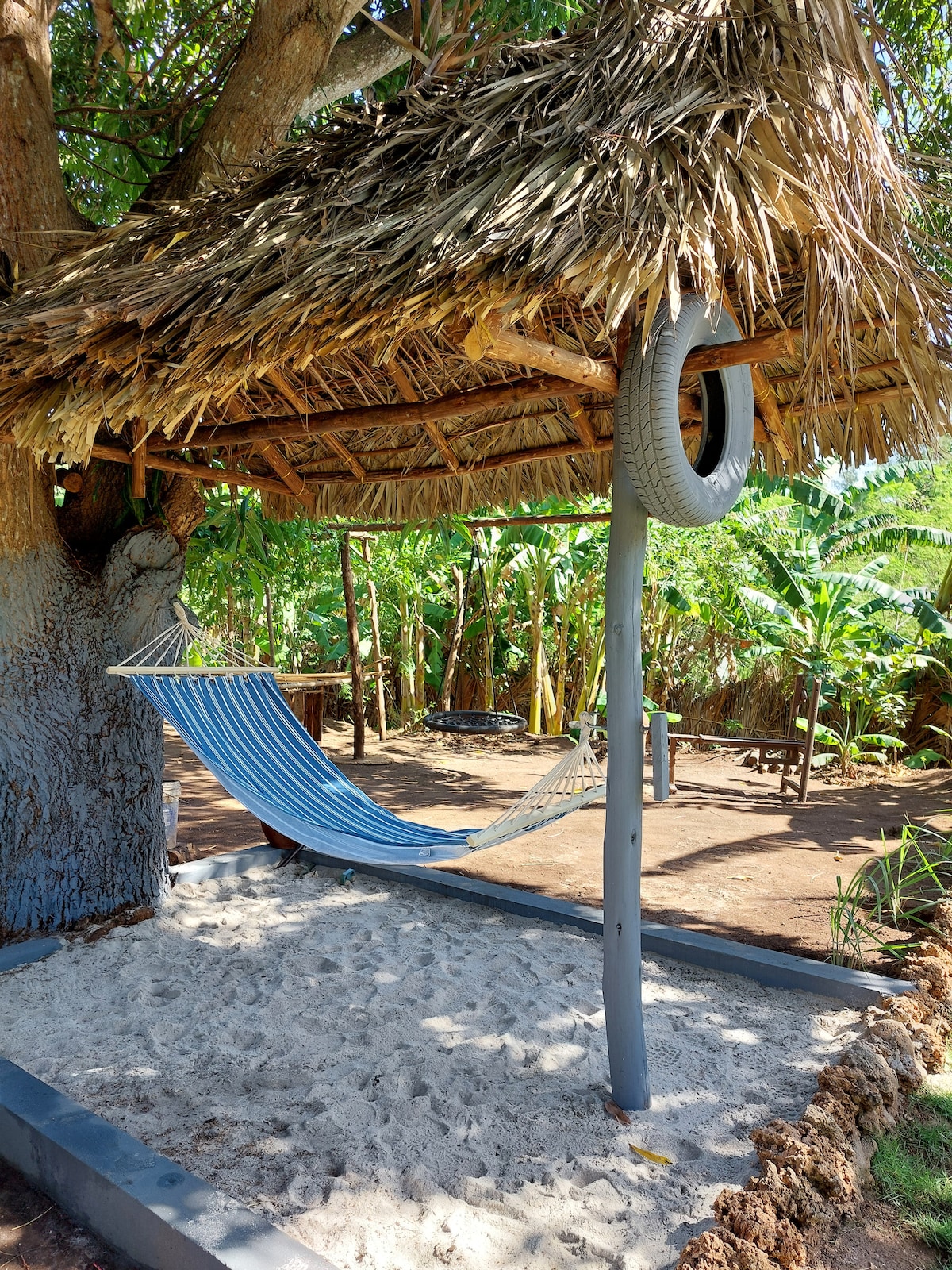 A shaded hammock is suspended beneath a thatched roof, offering a comfortable spot for relaxation. A sandy area surrounds the hammock, while lush greenery and trees create a secluded atmosphere, enhancing the serene outdoor space.