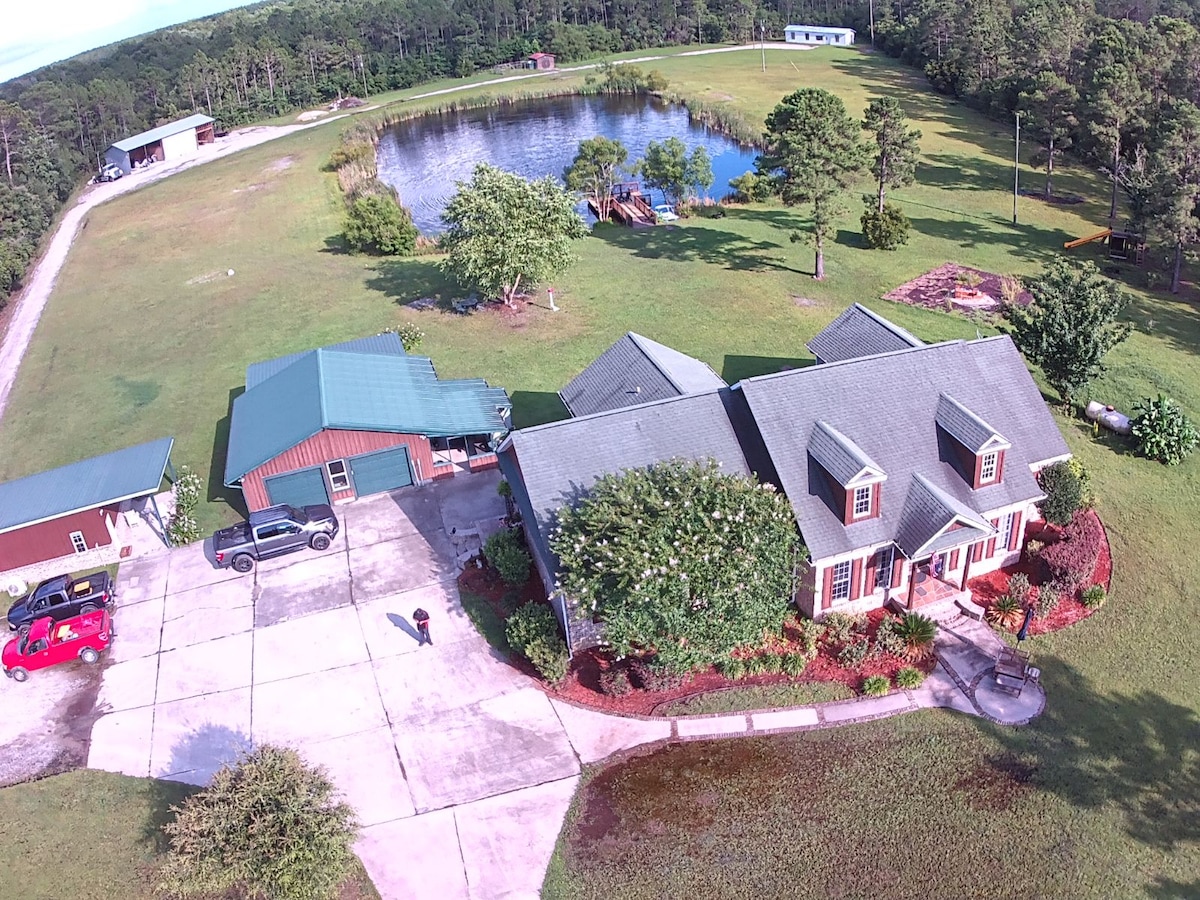 An aerial view showcases a spacious cabin with a green roof and a well-maintained exterior, surrounded by lush landscaping. A stocked pond is visible nearby, along with additional buildings and open grassy areas on the 8-acre estate.