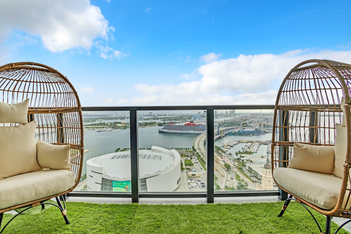Two stylish hanging chairs with soft cushions are positioned on a balcony, offering expansive views of the bay and marina. The surrounding area is graced with lush green grass, complementing the bright blue sky and clouds above.