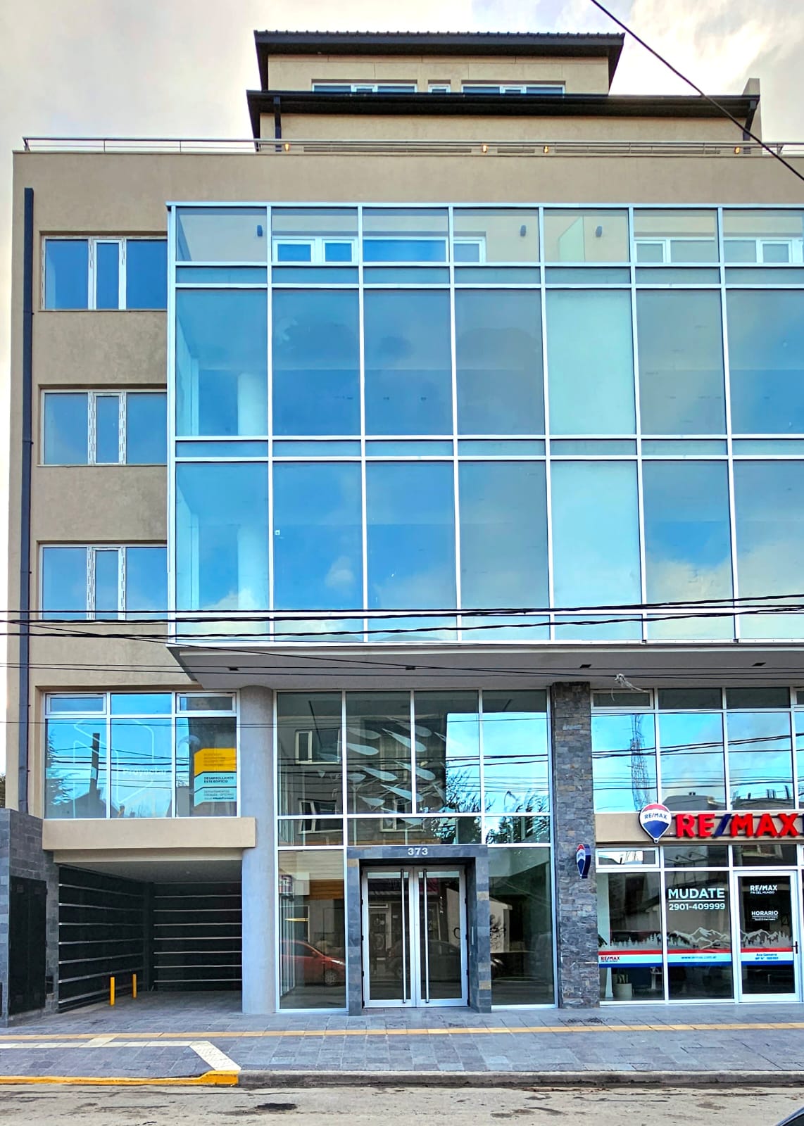 The exterior of a modern building is displayed, characterized by large glass windows that reflect the surrounding environment. A sleek entrance with an automatic sliding door is visible, along with a stone facade at the base and prominently placed signage.
