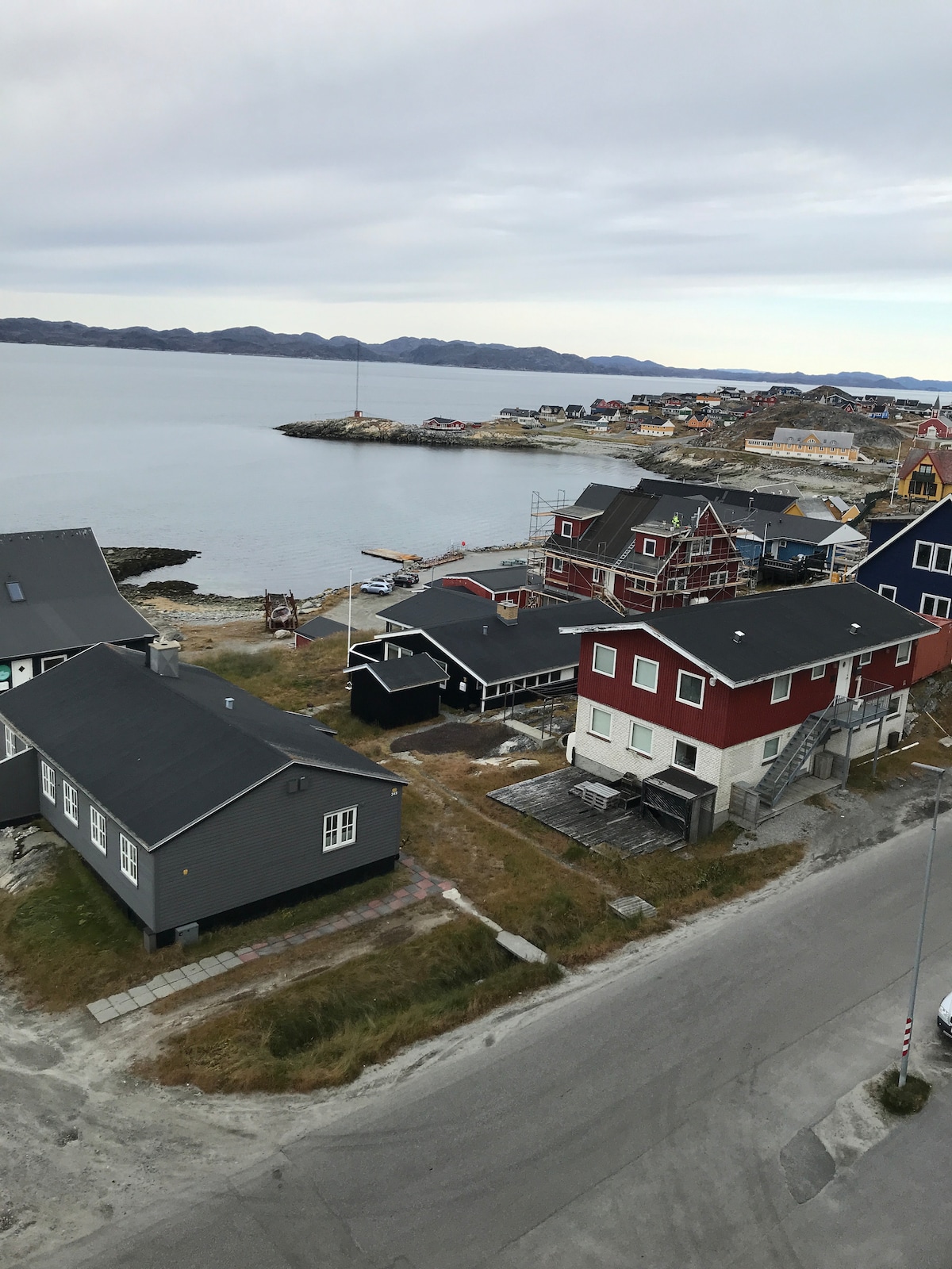 A panoramic view captures a coastal landscape with colorful houses lining the waterfront. The shoreline is dotted with boats, and distant hills provide a scenic backdrop. The surrounding area features a mix of buildings, highlighting a quiet, serene environment near the water.