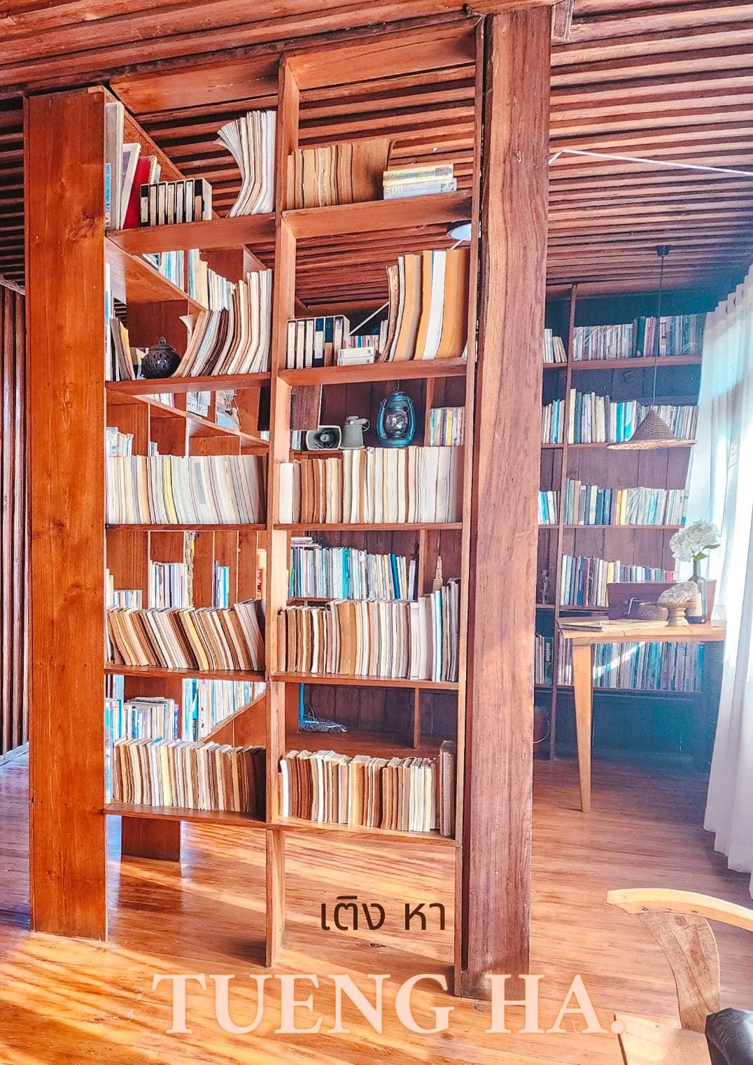 A wooden bookshelf filled with neatly organized books lines the walls of a cozy reading area. Soft sunlight filters through the nearby curtain, illuminating the space and drawing attention to a small table adorned with a decorative vase.