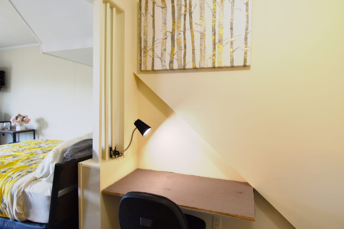 A work desk is positioned against the wall, illuminated by a small lamp. Above the desk, a piece of art featuring a forest scene adds a touch of nature. A cozy bed is partially visible in the background, draped with a yellow and gray comforter.