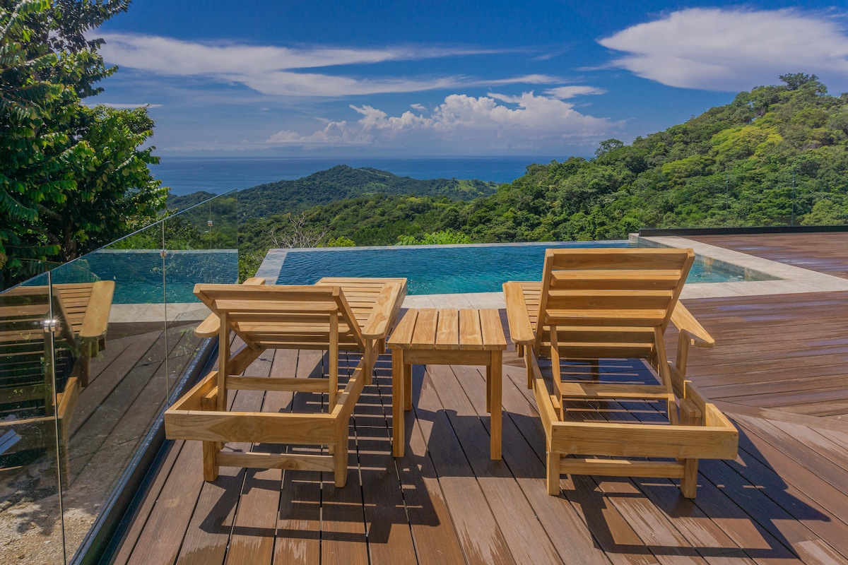 Two wooden lounge chairs accompany a small table on a spacious deck. A serene infinity pool extends beyond, blending into the surrounding lush greenery and scenic ocean views under a clear blue sky.