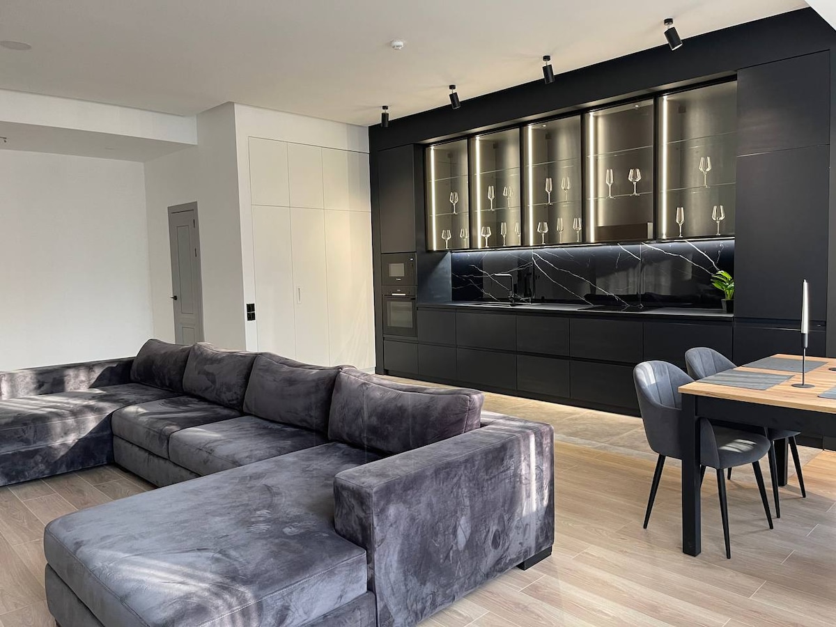 A modern living area features a spacious gray sectional sofa with clean lines. A sleek black kitchen with minimalist cabinetry runs along one wall, highlighted by elegant lighting. A wooden dining table accompanied by black chairs is positioned nearby, creating an inviting space for meals.
