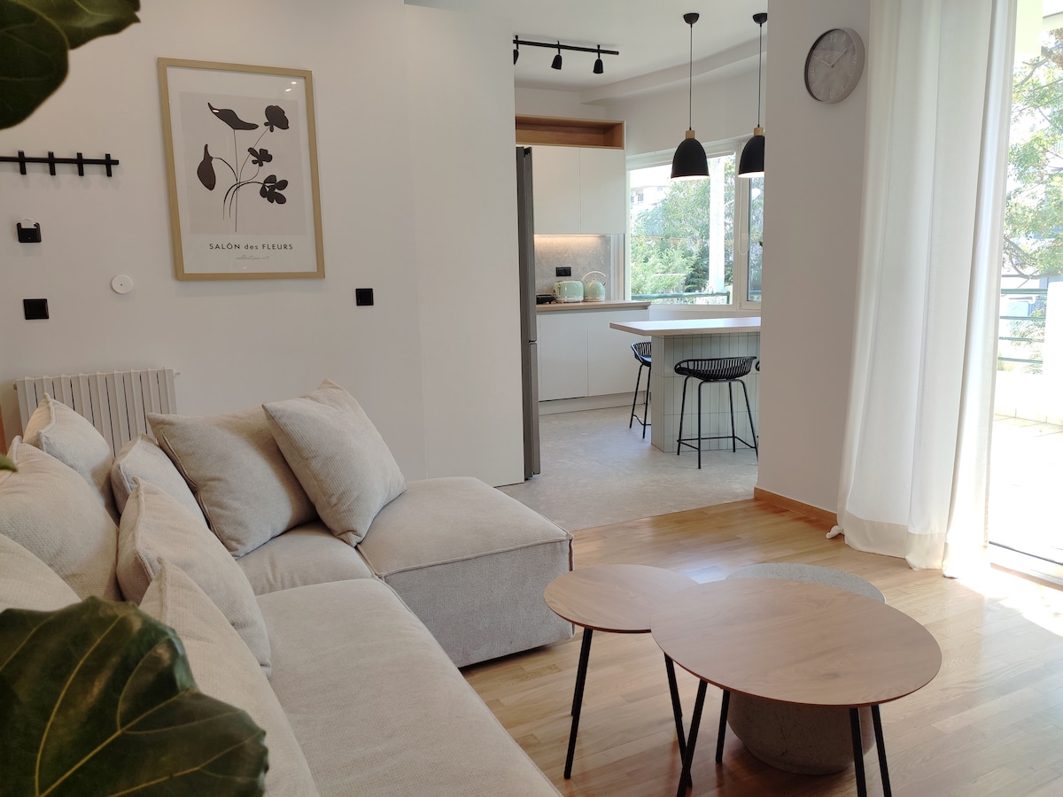 A modern living area features a light-colored sectional sofa arranged around a circular coffee table. Natural light fills the space through large windows, highlighting a minimalist kitchen with contemporary furnishings. Decorative plants enhance the inviting setting, complementing the neutral color palette.