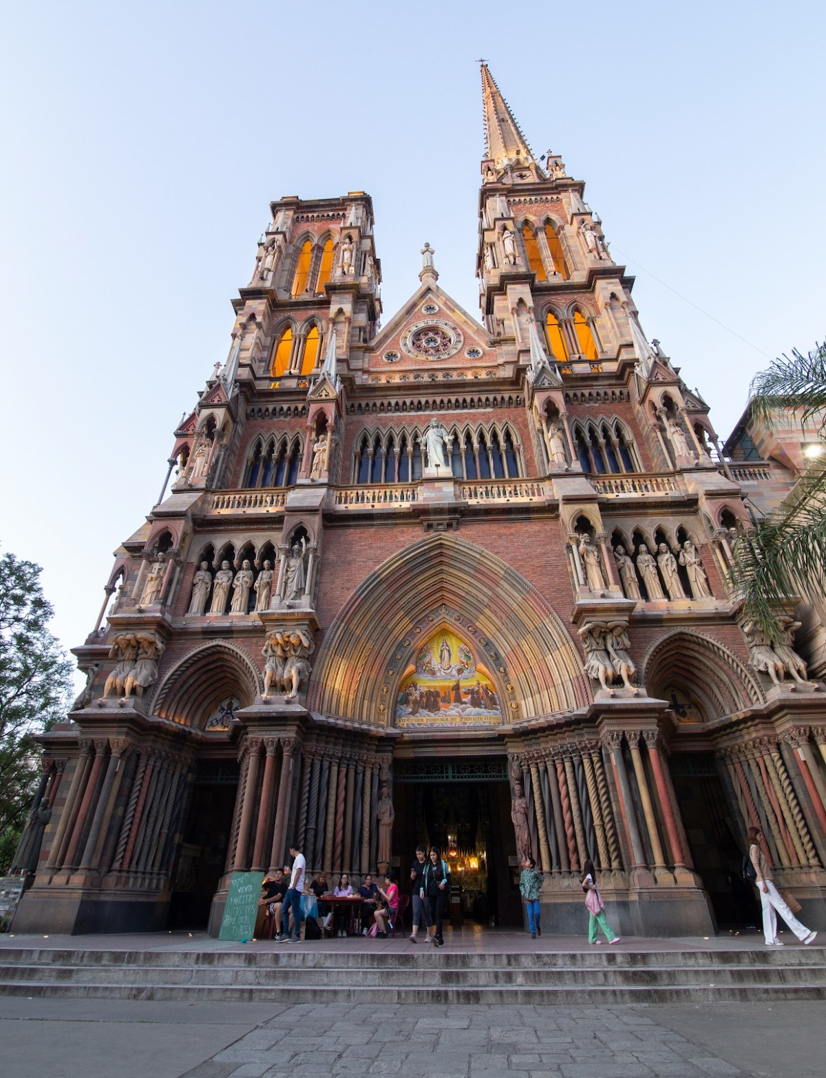 The exterior of a grand church features intricate brickwork and elaborate sculptures. Two tall spires rise above the main entrance, which is adorned with detailed carvings. Warm light spills from the windows as visitors gather outside, surrounded by lush greenery.