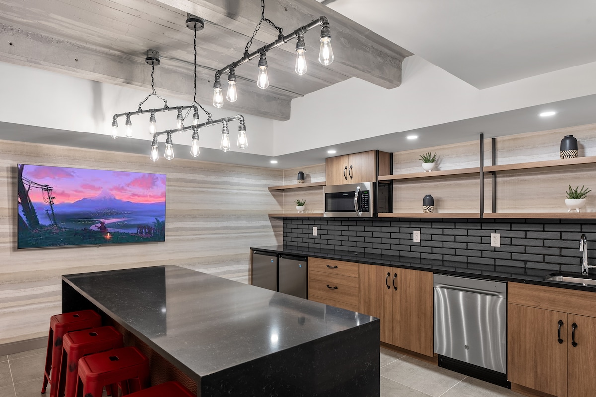 A modern kitchen area is presented with wooden cabinets and a dark countertops. Bright overhead lighting illuminates the space. Black subway tiles line the back wall. Seating is available at a large island with red stools, and a stainless steel dishwasher is integrated beneath the counter.