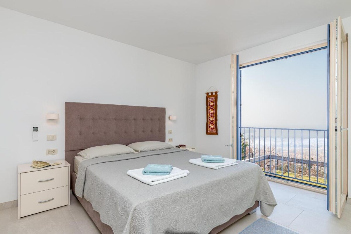 A spacious bedroom features a large bed dressed in soft linens. Natural light floods the room through open French doors, providing a view of the serene landscape outside. A bedside table stands next to the bed, adding functionality to the welcoming space.