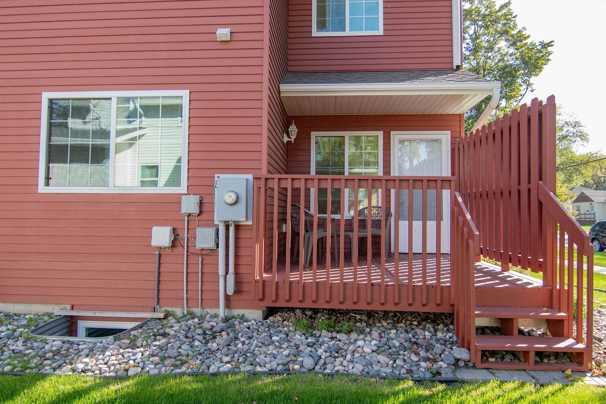 A wooden deck with a railing provides access to the townhome, featuring outdoor seating for enjoying the surroundings. The exterior showcases a deep red siding and several windows, while decorative gravel borders the foundation.