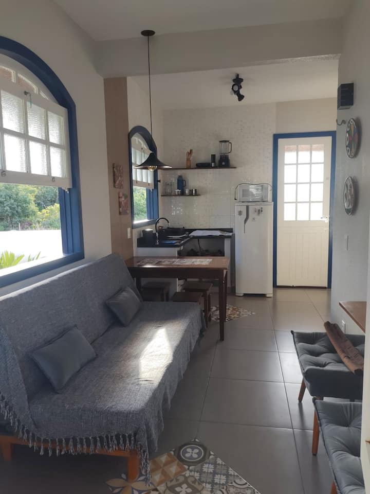 Sebule iliyo na kitanda cha sofa mara mbili, feni ya dari na televisheni ya inchi 50. Kwenye mandharinyuma, meza ya kulia chakula, jiko lenye vifaa.