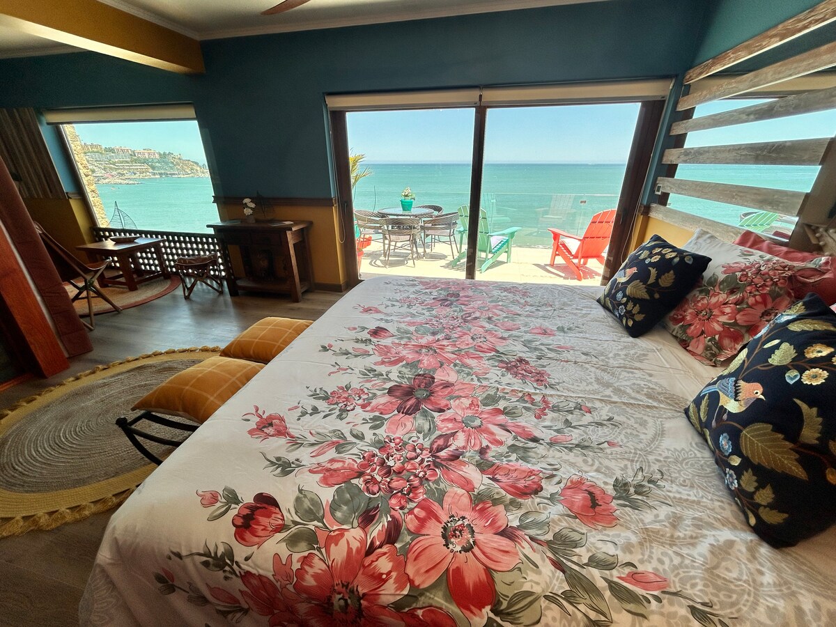 A spacious room features a brightly colored floral bedspread on a large bed, with decorative pillows. Large glass doors provide a stunning view of the ocean. Outdoor seating is visible on the terrace, allowing enjoyment of the coastal scenery.