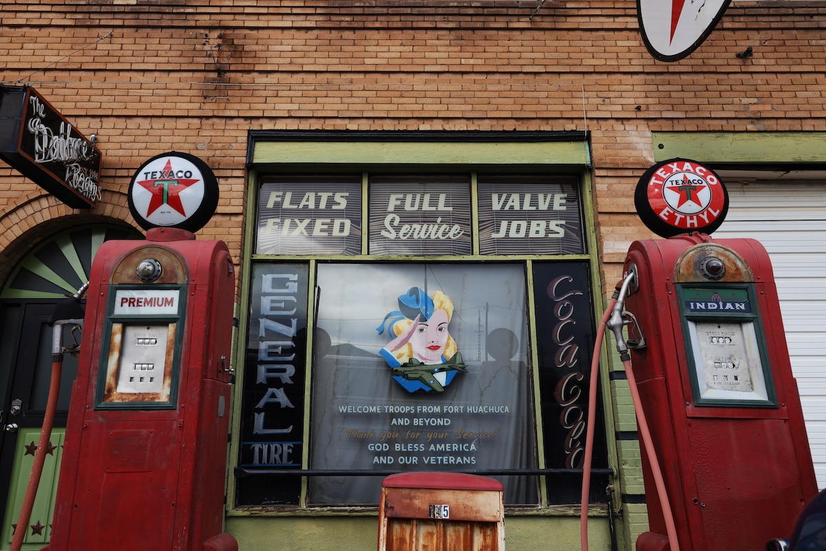 An old service station facade features vintage fuel pumps in red and white. A large sign displays the services offered, including 'FLATS FIXED,' 'FULL SERVICE,' and 'VALVE JOBS.' The colorful window decoration conveys a message of support for veterans and American troops.
