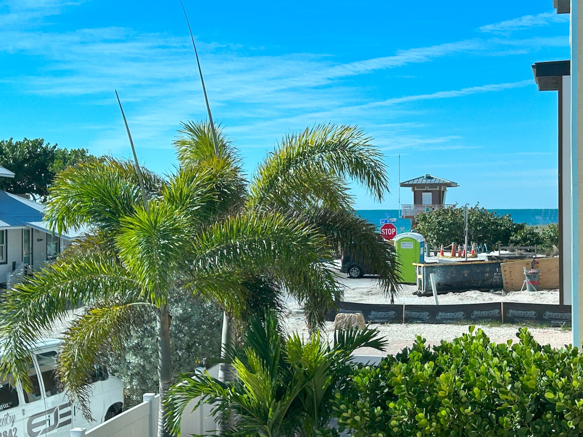 A view framed by vibrant palm trees and greenery shows a distant glimpse of the Gulf of Mexico. A sandy area is seen beyond the foliage, with a stop sign and a beach access point in the background under a clear blue sky.
