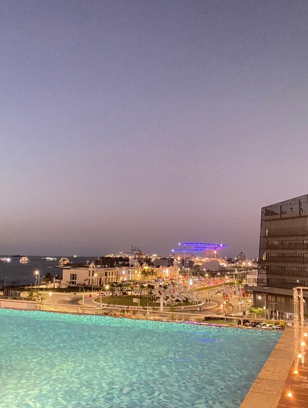 An infinity pool extends toward the horizon, with water reflecting the twilight sky. A vibrant cityscape is visible in the background, featuring illuminated buildings and greenery along a winding path, while boats can be seen on the water in the distance.
