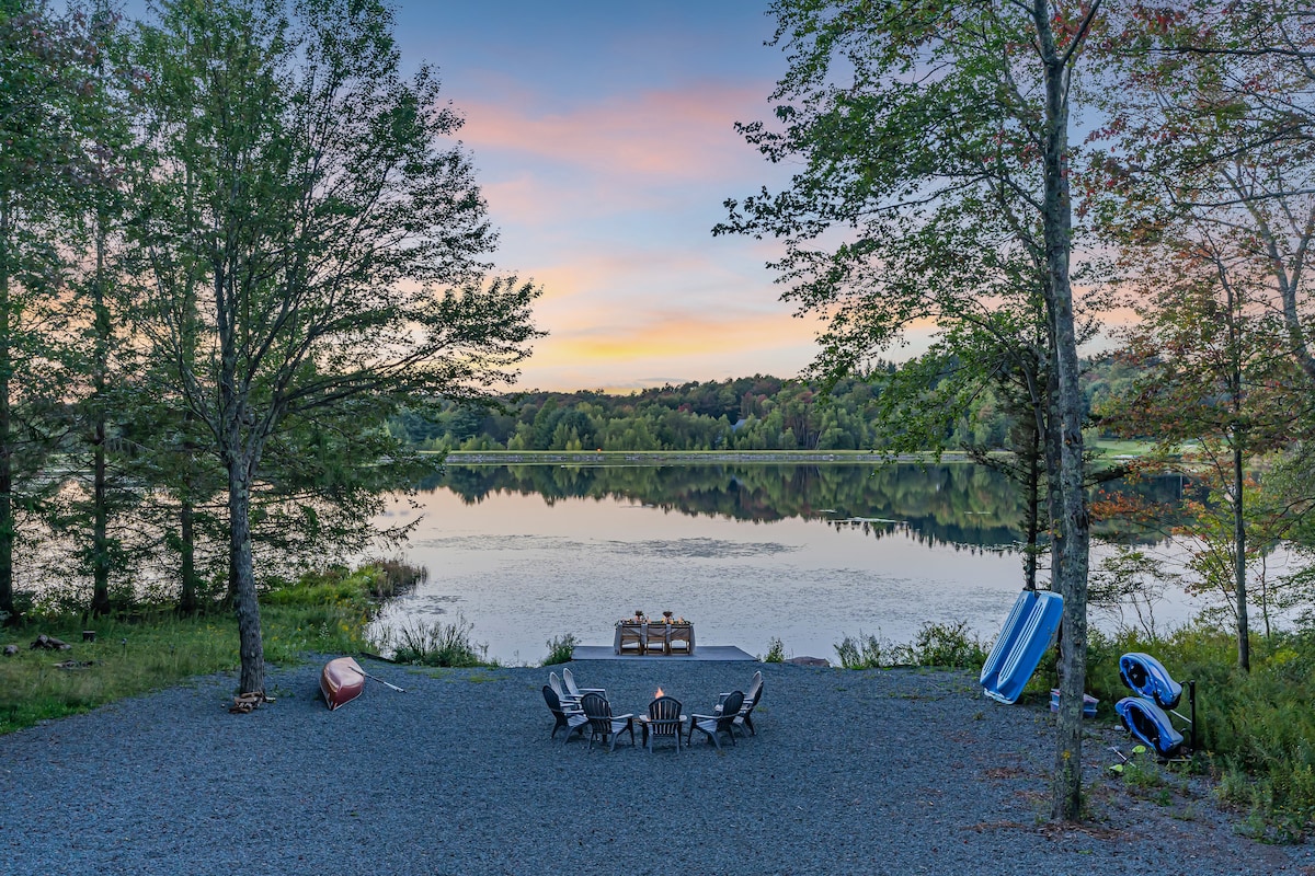 A gravel patio area near the lake features several outdoor chairs arranged around a fire pit. Kayaks and a canoe are positioned at the water's edge, while the tranquil lake reflects the colors of the sunset, framed by trees and gentle hillside in the background.