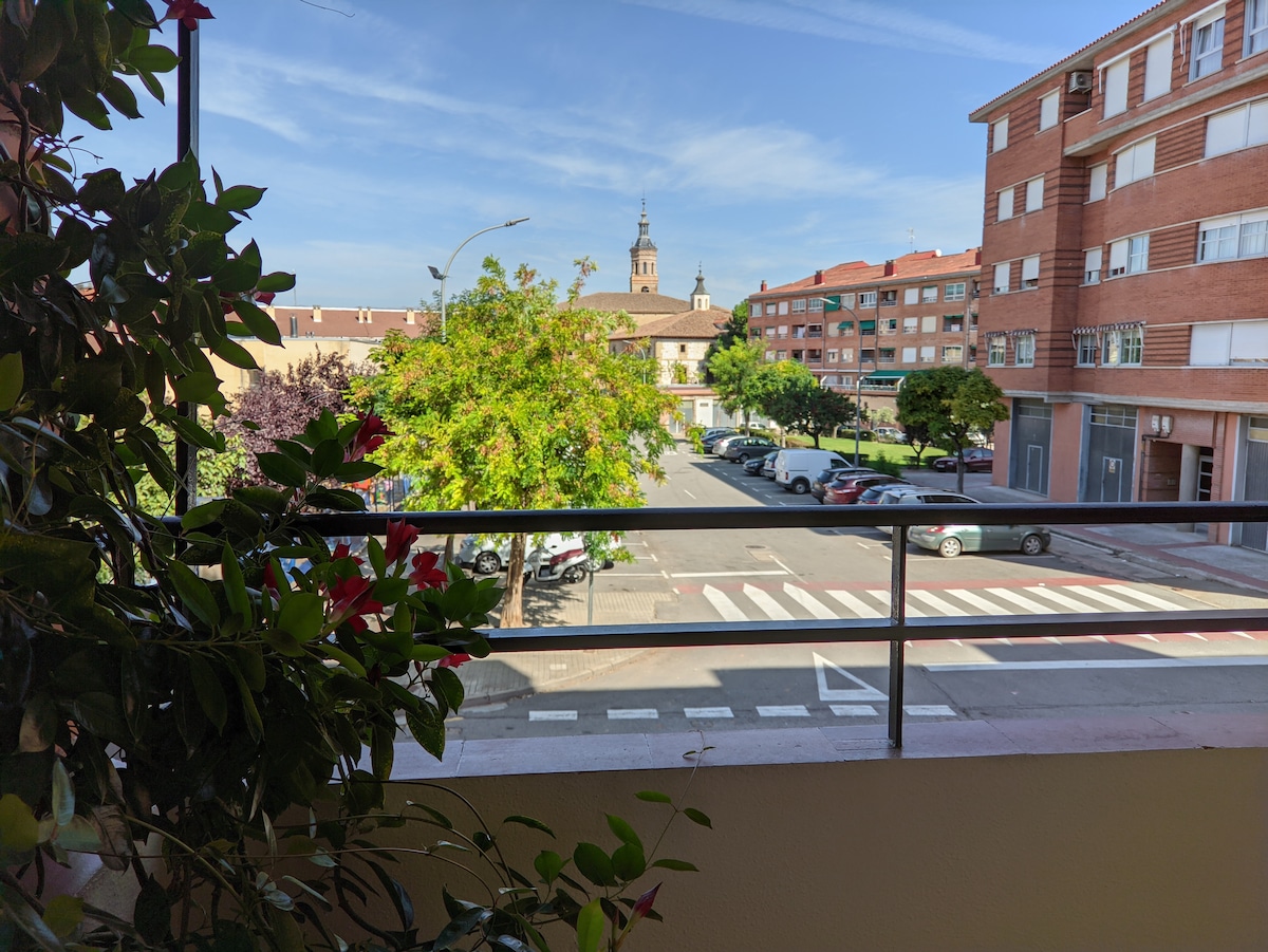 A view from a balcony overlooks a bustling street lined with parked cars. Lush greenery and colorful flowers frame the scene, while a clock tower in the distance adds a focal point to the charming neighborhood. Bright blue skies enhance the overall atmosphere.