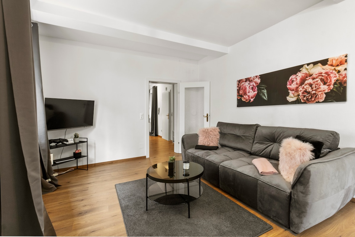 A cozy living area is furnished with a plush gray sofa and a round coffee table atop a soft rug. A wall-mounted television and floral artwork add character to the space. Natural light comes in through nearby windows, creating a welcoming atmosphere.