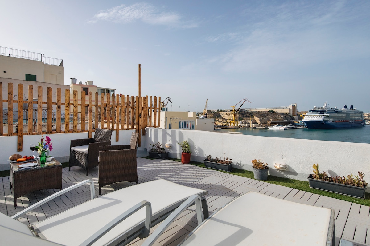 A spacious rooftop terrace is equipped with lounge chairs and comfortable seating, offering views of the shimmering water and nearby docked ships. Flower pots and greenery add a natural touch, while an inviting atmosphere is created by the open layout and panoramic surroundings.