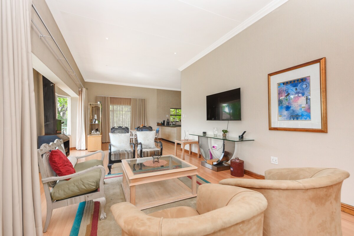 A living area features a comfortable seating arrangement with a mix of chairs and a sofa. A coffee table stands in the center, complemented by a wall-mounted television. Soft lighting enhances the neutral tones of the decor, while large windows provide views of the outside greenery.
