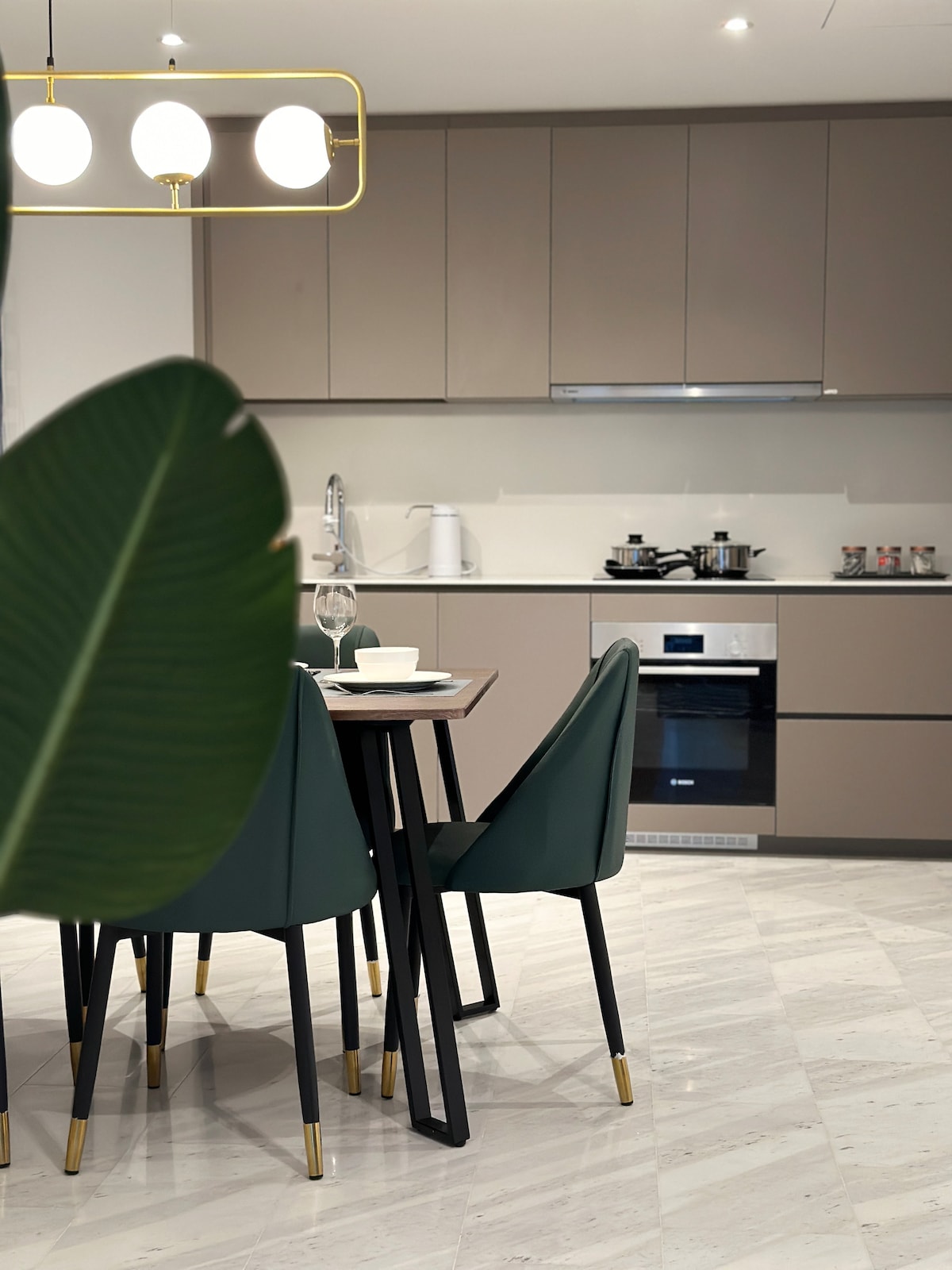 The kitchen area features modern cabinetry in neutral tones. A dining table with green chairs is set for meals. Appliances include an oven and a stovetop, complemented by countertop essentials discreetly arranged nearby. Natural light illuminates the marble flooring through a nearby window.