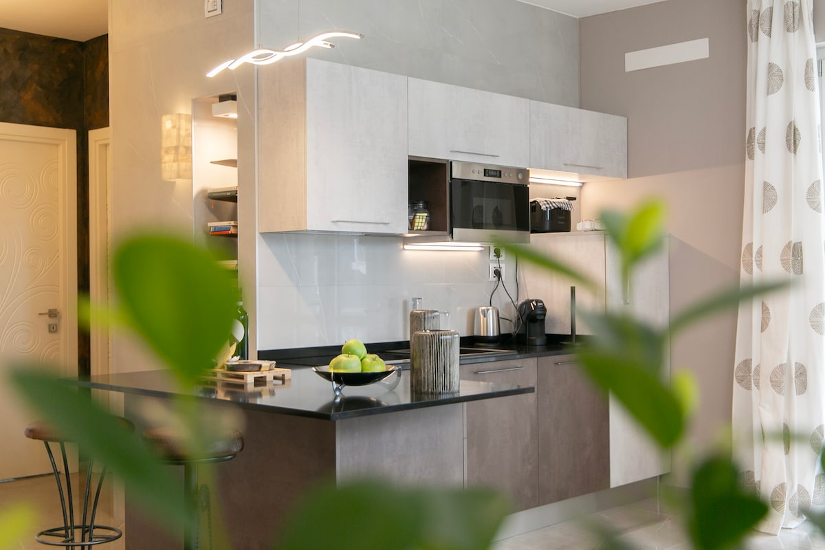 A modern kitchen area is visible, featuring sleek cabinetry and a dark countertop. A bowl of green apples sits atop the island, complemented by stainless steel appliances. Natural light filters in through a nearby window, partially obscured by decorative curtains.