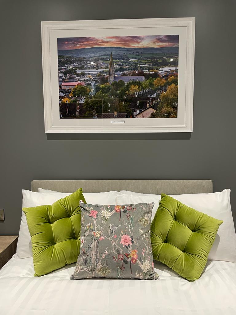 A cozy bed features a light-colored headboard and an arrangement of decorative pillows in shades of green and floral patterns. Above the bed, a framed photograph depicts a landscape of the Strabane area during sunset, highlighting the local scenery.