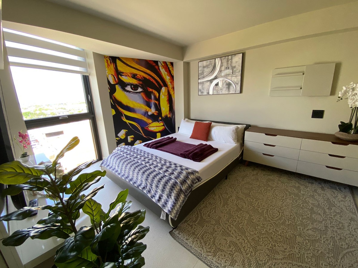 A stylish bedroom features a spacious bed adorned with decorative pillows and a soft throw blanket. A vibrant piece of artwork adds a pop of color to the wall, while a large window offers ample natural light. A dresser and a decorative plant complete the inviting space.