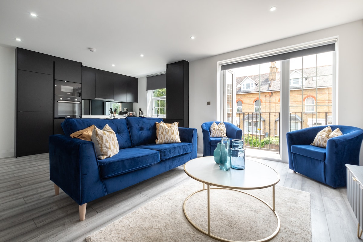 A modern living area features a spacious blue sofa with decorative cushions, complemented by two cozy armchairs. A round coffee table sits in front, and large glass doors provide access to a balcony, allowing natural light to fill the space.