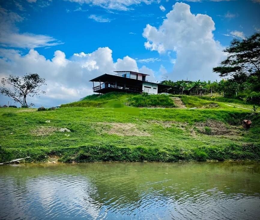 A large house sits on a hillside overlooking a calm body of water. Lush green grass surrounds the property, and a few trees are visible in the background under a partly cloudy sky. The house features a covered balcony that enhances outdoor relaxation.
