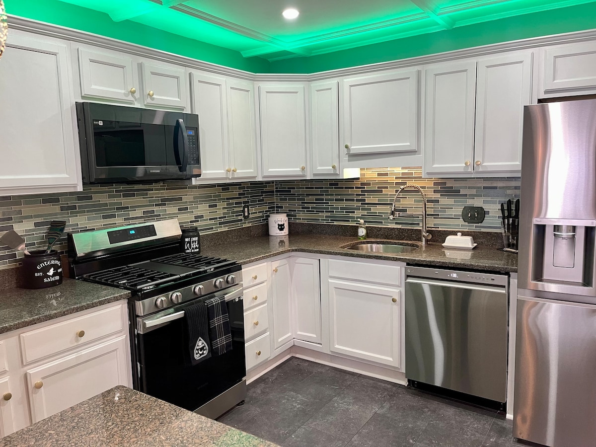 A modern kitchen features white cabinetry and stainless steel appliances, including a microwave, gas stove, and refrigerator. A sleek, stone countertop provides ample workspace, while mosaic tile backsplashes are illuminated by soft green lighting, adding a contemporary touch to the culinary area.