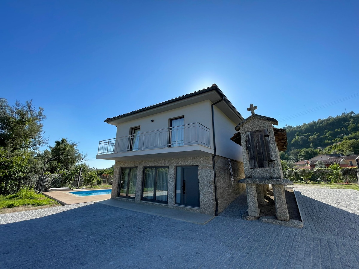A modern two-story house showcases a clean design, with large glass doors on the ground floor leading to an outdoor space. A traditional stone structure stands beside the house, highlighting the blend of contemporary and classic architecture. Clear skies and greenery enhance the serene setting.