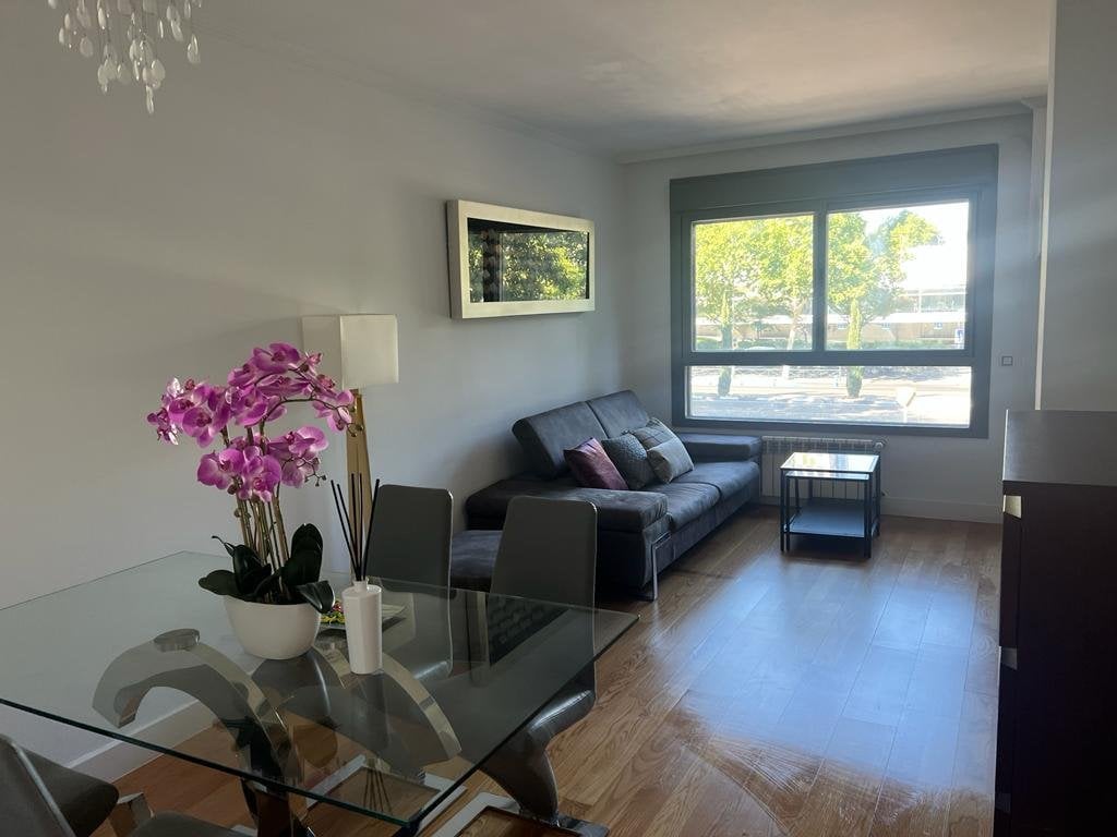 An open living area presents a comfortable sofa adorned with decorative cushions and a coffee table. A dining table with modern chairs is set nearby. Natural light streams through large windows showcasing views of outside greenery, enhancing the space's bright atmosphere.