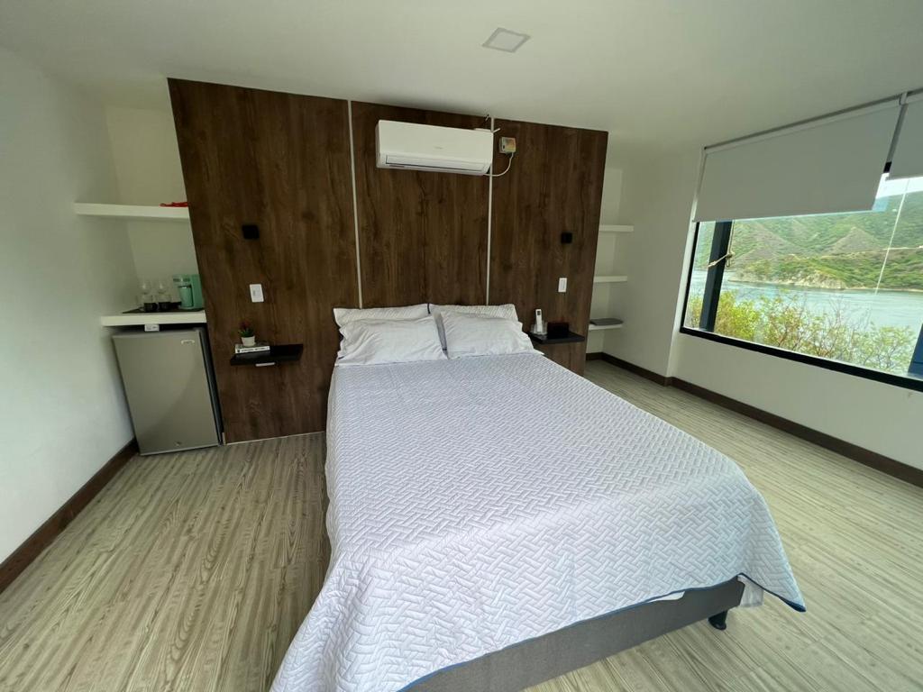 A modern room features a spacious double bed covered with a light blanket. Shelves are mounted on the walls, and a small minibar is positioned near the bed. A large window offers views of the surrounding greenery and water.